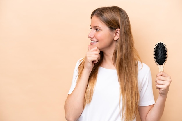 Junge hübsche blonde Frau mit Haarkamm isoliert auf beigem Hintergrund, denkt an eine Idee und schaut zur Seite