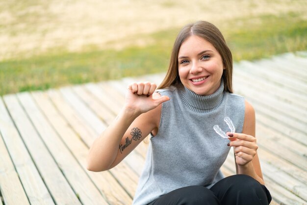 Junge hübsche blonde Frau mit unsichtbaren Zahnspangen im Freien stolz und selbstzufrieden