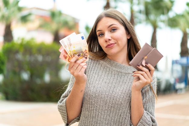 Junge hübsche Blondine, die Geldbörse mit Geld hält und Zweifel hat