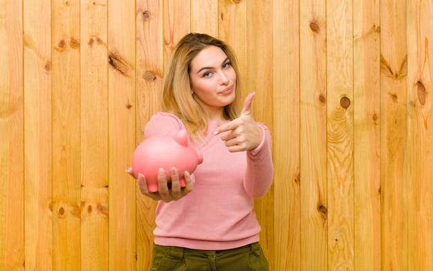 Junge hübsche Blondine mit einem Sparschwein auf hölzerner Wand