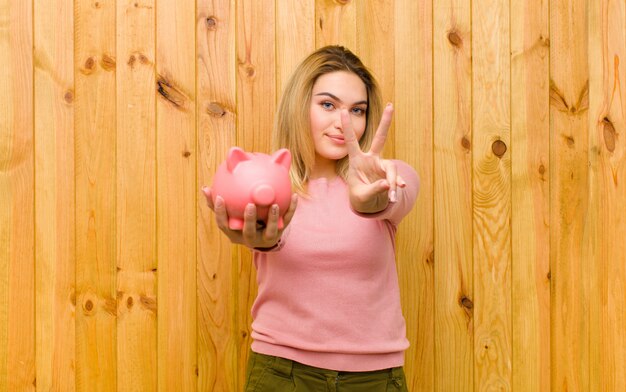 Junge hübsche Blondine mit einem Sparschwein gegen hölzerne Wand