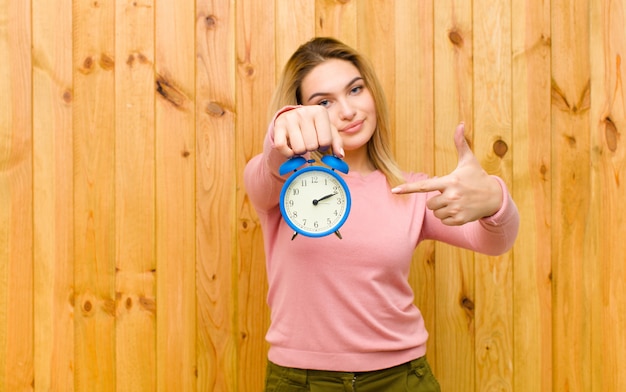 Junge hübsche Blondine mit einem Wecker auf hölzerner Wand