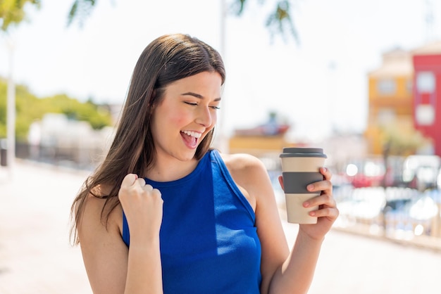 Junge hübsche Brasilianerin hält draußen einen Kaffee zum Mitnehmen in der Hand und feiert einen Sieg