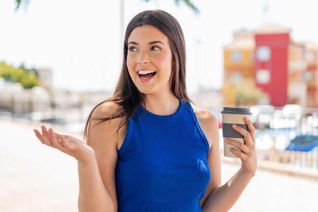 Junge hübsche Brasilianerin hält mit überraschendem Gesichtsausdruck im Freien einen Kaffee zum Mitnehmen in der Hand