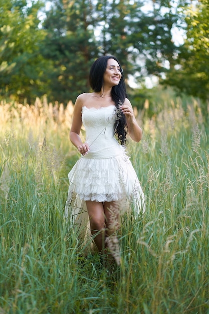 Junge hübsche Brunettefrau im weißen Hochzeitskleid, gehend in den Wald und lächeln