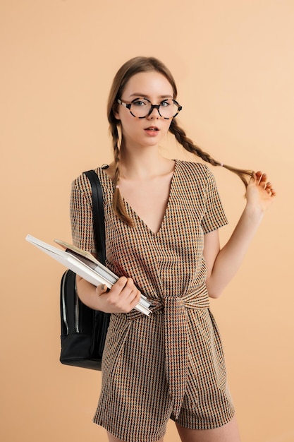 Junge hübsche Dame mit zwei Zöpfen in Tweedoverall und Brille mit schwarzem Rucksack auf der Schulter, die Buch und Notizblöcke in der Hand hält, während sie verträumt in die Kamera über beigen Hintergrund schaut