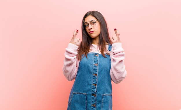 Junge hübsche Frau, die ängstlich die Finger kreuzt und auf Glück mit einem besorgten Blick gegen rosa Wand hofft.