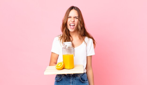 Junge hübsche Frau, die aggressiv schreit, sehr wütend aussieht und ein Tablett mit Orangensaft hält