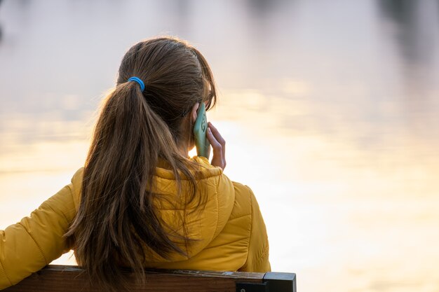 Junge hübsche Frau, die auf einer Parkbank sitzt und draußen auf ihrem Smartphone spricht.