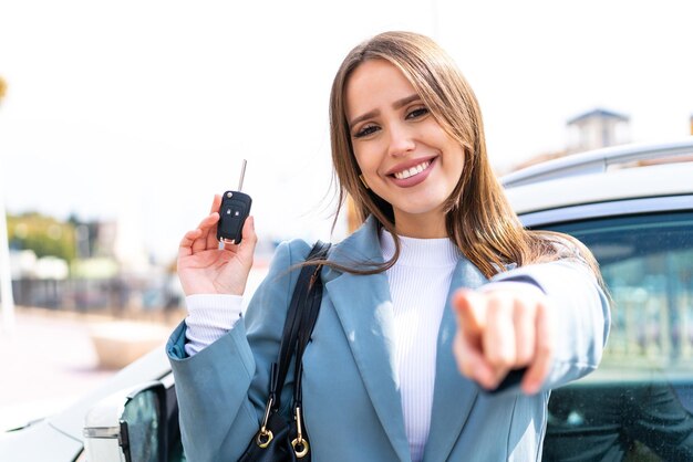 Junge hübsche Frau, die Autoschlüssel im Freien hält, zeigt mit einem selbstbewussten Ausdruck mit dem Finger auf Sie