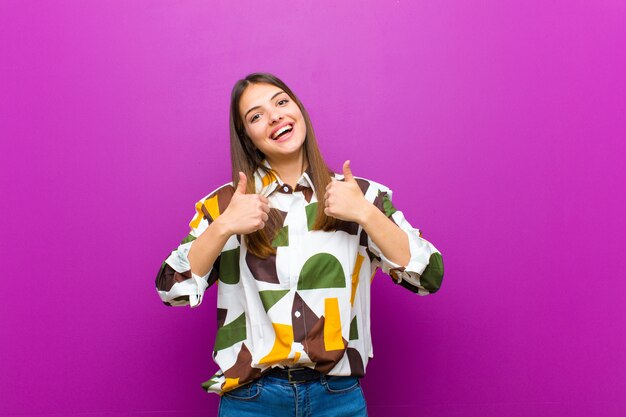 Junge hübsche Frau, die breit glücklich, positiv, selbstbewusst und erfolgreich lächelnd aussieht, mit beiden Daumen gegen lila Wand