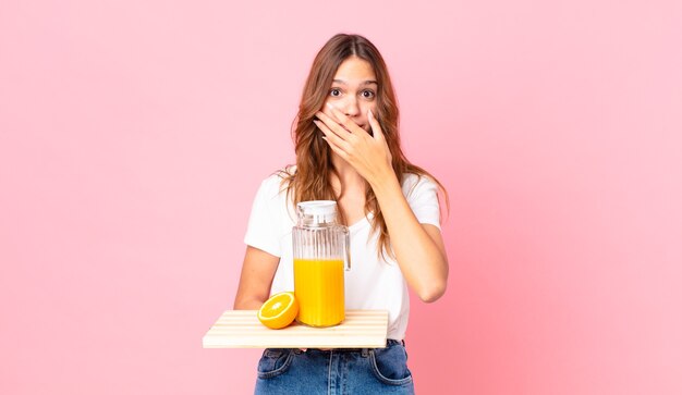 Junge hübsche Frau, die den Mund mit den Händen mit einem schockierten bedeckt und ein Tablett mit einem Orangensaft hält