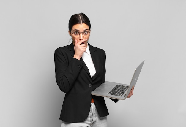 Junge hübsche Frau, die den Mund mit den Händen mit einem schockierten, überraschten Ausdruck bedeckt, ein Geheimnis hält oder oops sagt. Laptop-Konzept