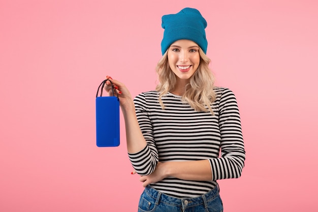 Junge hübsche Frau, die drahtlosen Lautsprecher hält, der Musik trägt, die gestreiftes Hemd und blauen Hut trägt, der glückliche positive Stimmung lächelt, die auf rosa Wand isoliert aufwirft