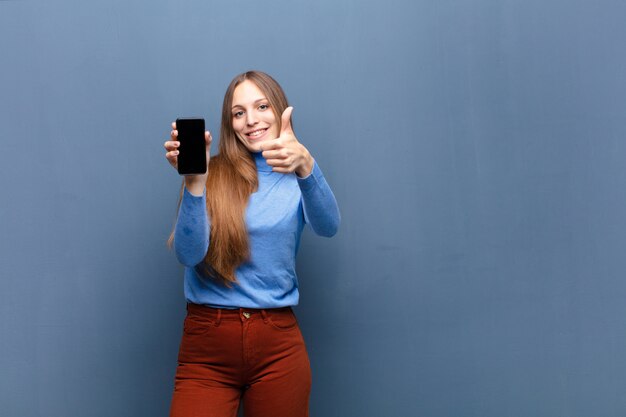 Junge hübsche Frau, die ein intelligentes Telefon gegen blaue Wand mit einem Kopienraum verwendet