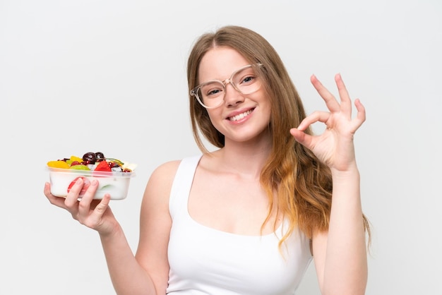 Junge hübsche Frau, die eine Schale mit Obst isoliert auf weißem Hintergrund hält und mit den Fingern ein Ok-Zeichen zeigt