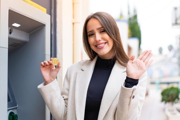 Junge hübsche Frau, die einen Bitcoin im Freien hält und mit der Hand mit glücklichem Ausdruck salutiert