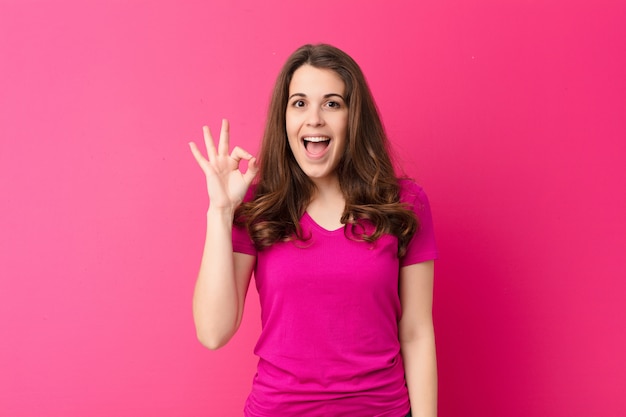 Junge hübsche Frau, die erfolgreich und zufrieden fühlt, mit weit geöffnetem Mund lächelt und okay Zeichen mit Hand über rosa Wand macht