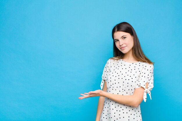 Junge hübsche Frau, die fröhlich lächelt, sich glücklich fühlt und ein Konzept im Kopienraum mit Handfläche gegen blaue Wand zeigt
