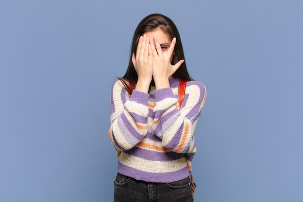 junge hübsche frau, die gesicht mit den händen bedeckt, mit überraschtem ausdruck zwischen den fingern späht und zur seite schaut. studentisches Konzept