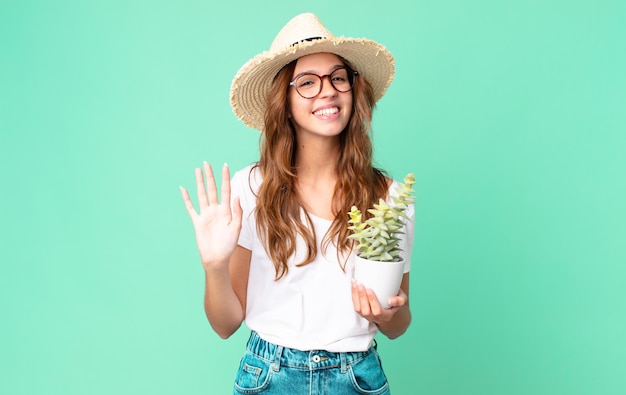 Foto junge hübsche frau, die glücklich lächelt, mit der hand winkt, sie mit einem strohhut begrüßt und begrüßt und einen kaktus hält