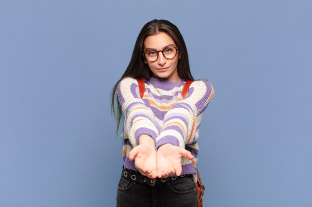 Junge hübsche Frau, die glücklich mit freundlichem, selbstbewusstem, positivem Blick lächelt, ein Objekt anbietet und zeigt