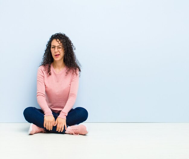 Junge hübsche Frau, die glücklich und freundlich schaut, lächelt und ein Auge auf Sie mit einer positiven Haltung zwinkert, die auf Boden sitzt