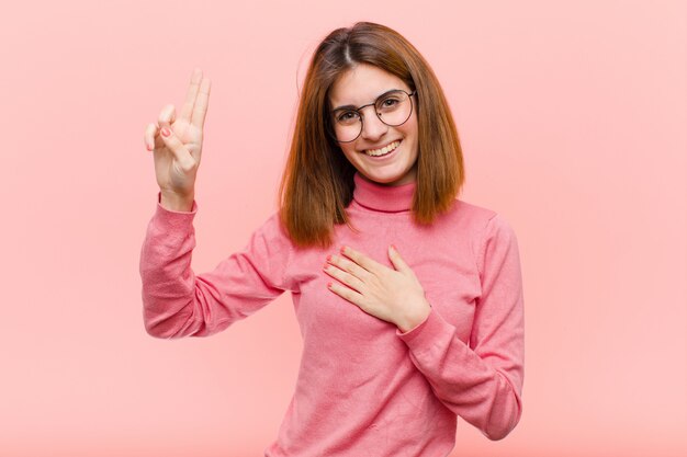 Junge hübsche Frau, die glücklich, zuversichtlich und vertrauenswürdig aussieht, lächelt und Siegeszeichen zeigt, mit einer positiven Haltung über rosa Wand