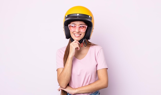Junge hübsche Frau, die mit einem glücklichen, selbstbewussten Ausdruck mit der Hand am Kinn lächelt. Motorradfahrer und Helm