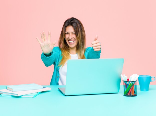 Junge hübsche Frau, die mit einem Laptop lächelt und schaut freundlich arbeitet und zeigt Nr. Sechs oder Sechstel mit der Hand vorwärts und zählt unten