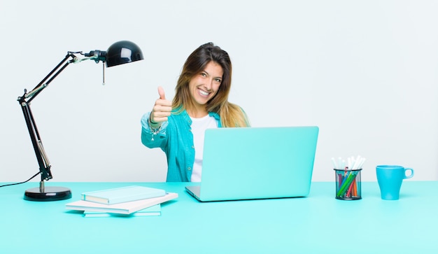 Junge hübsche Frau, die mit einem Laptop sich fühlt stolz, sorglos, überzeugt und glücklich, positiv lächelnd mit den Daumen oben arbeitet