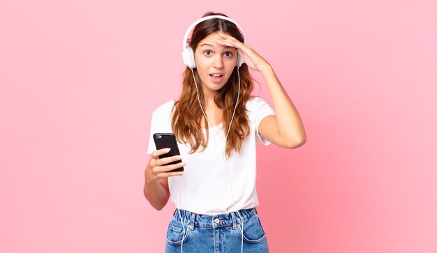 Junge hübsche Frau, die mit Kopfhörern und einem Smartphone glücklich, erstaunt und überrascht aussieht
