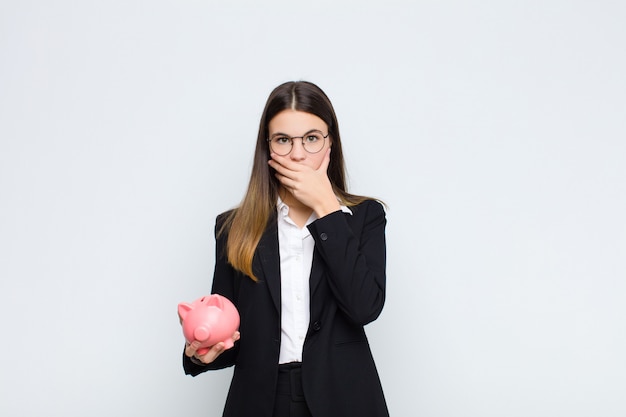 Junge hübsche Frau, die Mund mit den Händen mit einem schockierten, überraschten Ausdruck bedeckt, ein Geheimnis hält oder mit einem Sparschwein oops sagt