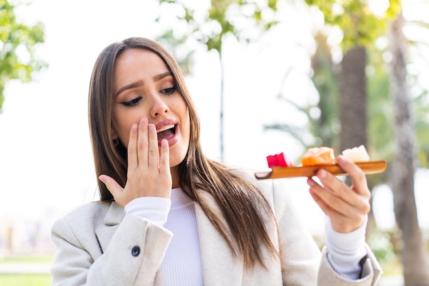 Junge hübsche Frau, die Sashimi im Freien mit Überraschung und schockiertem Gesichtsausdruck hält