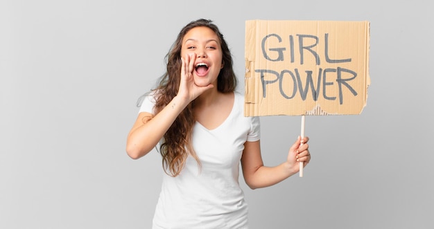 Junge hübsche Frau, die sich glücklich fühlt, mit den Händen neben dem Mund einen großen Schrei ausspricht und ein Girl-Power-Banner hält