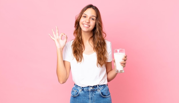 Junge hübsche Frau, die sich glücklich fühlt, Zustimmung mit einer okayen Geste zeigt und ein Glas Milch hält