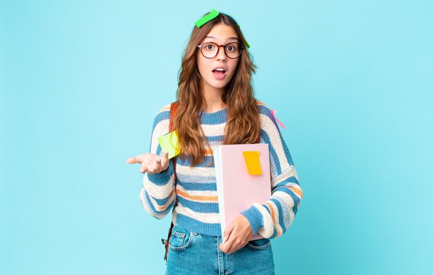 Junge hübsche Frau, die sich mit einer Tasche extrem schockiert und überrascht fühlt und Bücher hält