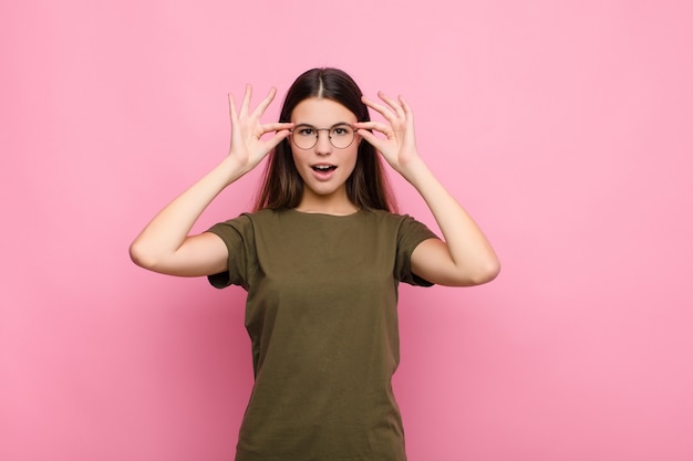 Junge hübsche Frau, die sich schockiert, erstaunt und überrascht fühlt und eine Brille mit erstauntem, ungläubigem Blick gegen die rosa Wand hält