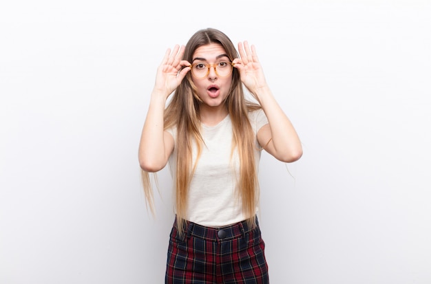 Junge hübsche Frau, die sich schockiert, erstaunt und überrascht fühlt und eine Brille mit erstauntem, ungläubigem Blick gegen die weiße Wand hält