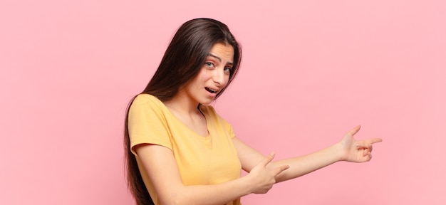 Junge hübsche frau, die sich schockiert und überrascht fühlt und mit erstauntem, offenem blick auf den kopienraum auf der seite zeigt