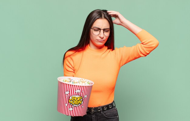 Junge hübsche Frau, die sich verwirrt und verwirrt fühlt, den Kopf kratzt und zur Seite schaut. Popcorn-Konzept