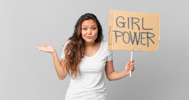 Junge hübsche Frau, die sich verwirrt und verwirrt fühlt und zweifelt und ein Girl-Power-Banner hält