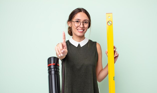 Junge hübsche Frau, die stolz und selbstbewusst lächelt und die Nummer eins macht. Architekturkonzept