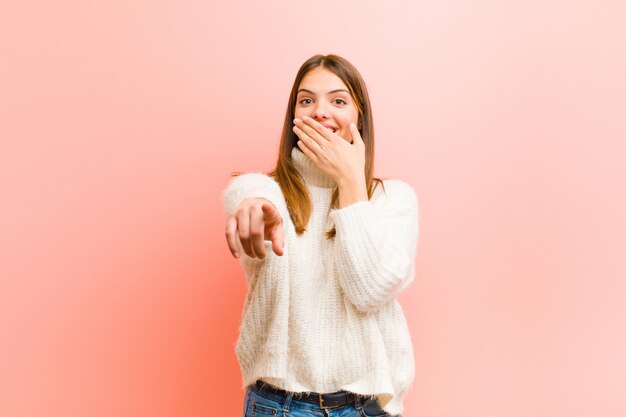 Junge hübsche Frau, die über Sie lacht, zur Kamera zeigt und sich über Sie lustig macht oder Sie gegen rosa Wand verspottet