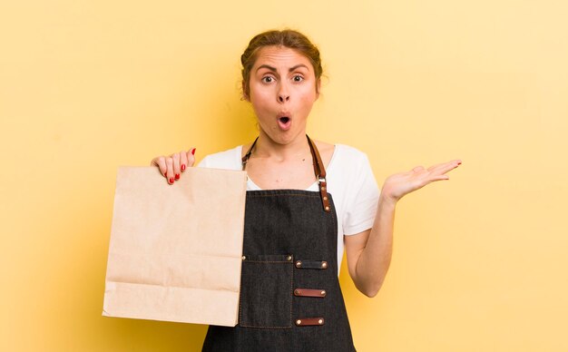 Junge hübsche Frau, die überrascht und schockiert aussieht, mit heruntergefallenem Kiefer, die ein Objekt-Fast-Food-Kurierkonzept hält