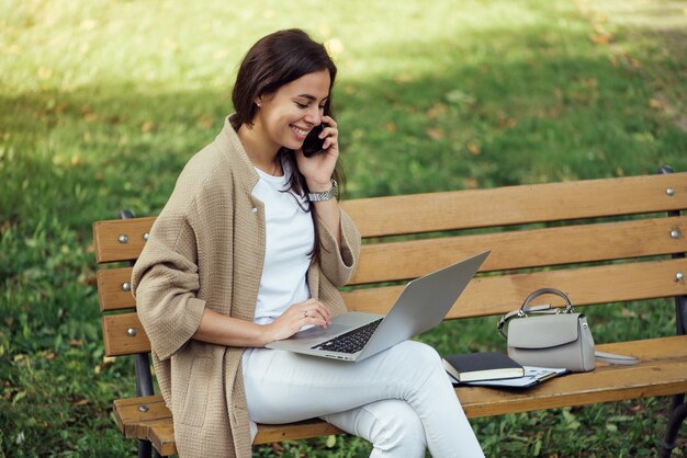 Junge hübsche Frau, die während des Sommertages auf einer Bank im Park sitzt und Laptop benutzt