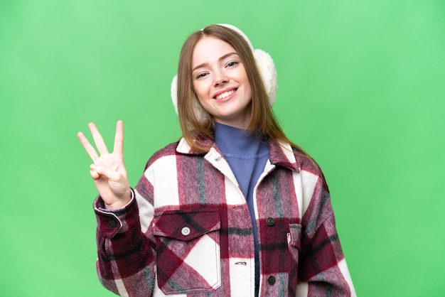 Junge hübsche Frau, die Wintermuffs über isoliertem Chroma-Key-Hintergrund trägt, glücklich und drei mit den Fingern zählend