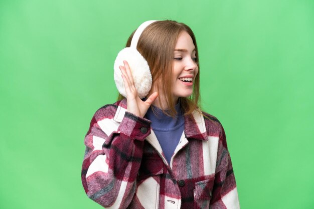 Junge hübsche Frau, die Wintermuffs über isoliertem Chroma-Key-Hintergrund trägt und etwas hört, indem sie die Hand auf das Ohr legt