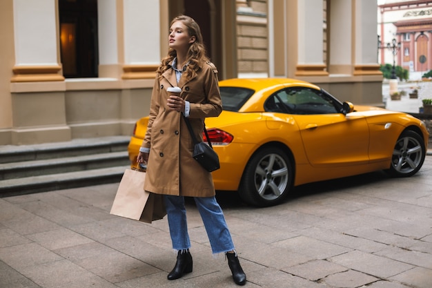 Junge hübsche Frau im Trenchcoat mit der kleinen schwarzen Kreuztasche, die Tasse Kaffee hält, um zu gehen und Einkaufstaschen in Händen, während nachdenklich