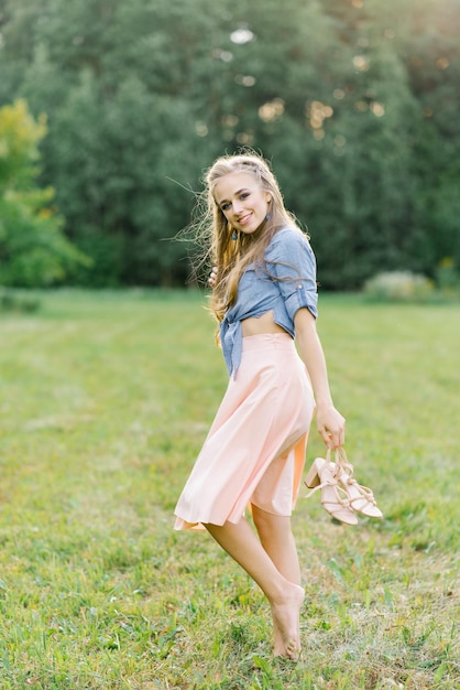 Junge hübsche Frau in einem Sommerkleid geht durch eine Wiese auf grünem Gras Ein junges Mädchen hält Schuhe in ihren Händen, die ihr schönes Bein zeigen Sommererholung im Freien